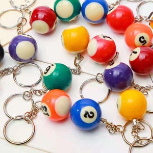 May include: A selection of billiard ball keyrings. Each keyring has a small pool ball with a number, connected to a silver-coloured metal ring. The balls are in various colours, including red, blue, green, and yellow.