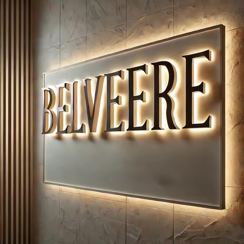 May include: A rectangular wall-mounted sign featuring the word BELVEERE in large, dark, three-dimensional capital letters. The sign is illuminated from behind, creating a soft, warm glow that highlights the edges of the lettering and the panel.
