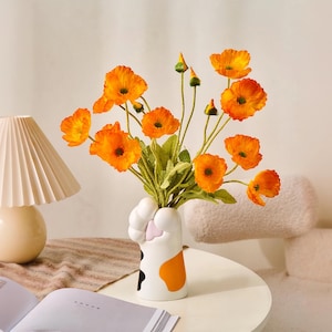 Puede incluir: Un jarrón de cerámica blanca con forma de pata de gato contiene un ramo de flores de color naranja brillante. El jarrón tiene marcas negras y naranjas. También se ven una lámpara color crema y un libro abierto.