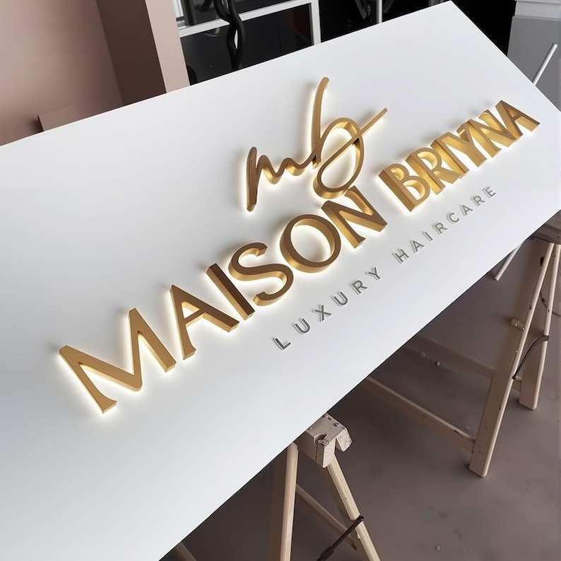 May include: A white rectangular sign featuring three-dimensional gold lettering that reads MAISON BRIYNA with a stylized mb logo above and the text LUXURY HAIRCARE in smaller gold letters below. The sign is mounted on a wooden easel stand.