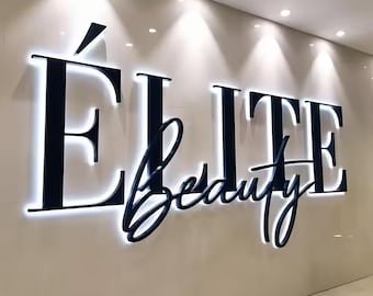 May include: A wall-mounted sign with the words "Élite Beauty" in a dark blue, elegant font. The letters are illuminated with a white glow, creating a striking contrast against the light-colored wall. The sign is a decorative element.