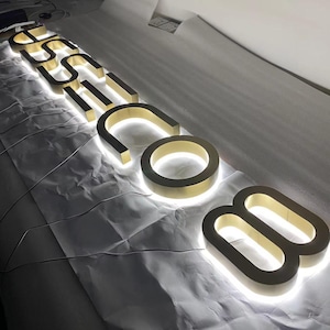 May include: A set of three-dimensional gold-colored metal letters spelling out the text "USSI.COM" with integrated white LED backlighting that creates a glowing halo effect around the edges of each character, arranged in a linear sequence.