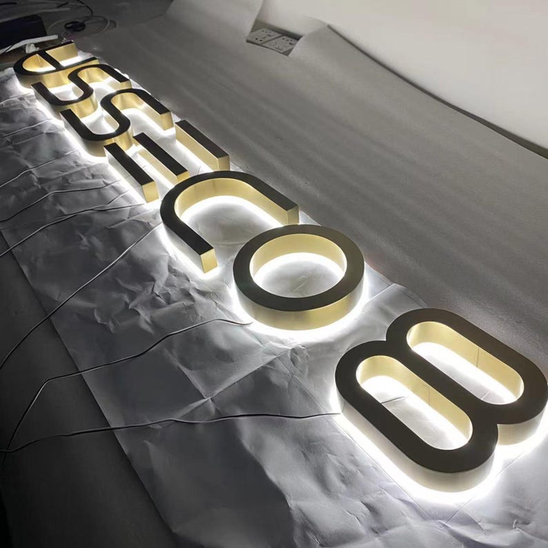 May include: A set of three-dimensional gold-colored metal letters spelling out the text "USSI.COM" with integrated white LED backlighting that creates a glowing halo effect around the edges of each character, arranged in a linear sequence.