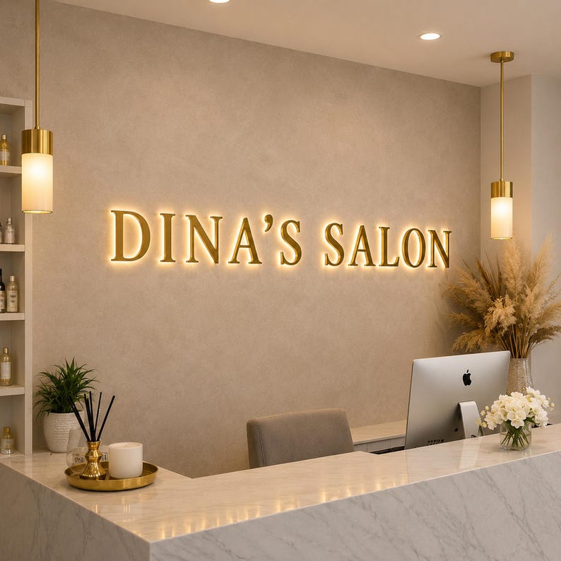 May include: A reception area featuring a wall-mounted sign that reads DINA'S SALON in illuminated gold lettering. The space includes a white marble counter, a computer monitor, two gold pendant lights, a potted plant, and a vase of white flowers.