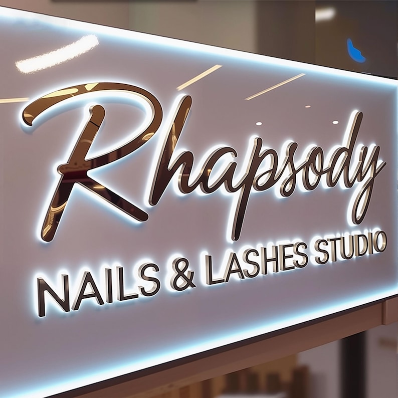 May include: A rectangular sign with a white background and a glowing blue light border. The sign features the word Rhapsody in a large, metallic, cursive font, followed by the text NAILS & LASHES STUDIO in a smaller, metallic, sans-serif font.