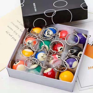 May include: A collection of billiard ball keyrings presented in a white box. Each keyring showcases a miniature pool ball with numbers and stripes, connected to a silver ring. The box is black with white circles and the text "MADE IN CHINA".