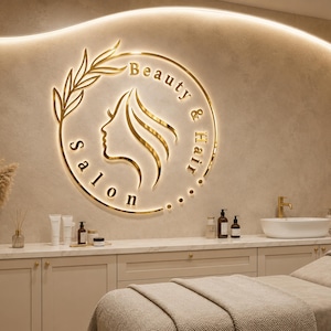 May include: A gold-colored circular wall sign featuring a profile silhouette of a face and the text Beauty & Hair Salon. The sign is illuminated with backlighting and mounted on a beige wall above a white counter with various salon supplies.