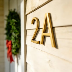 May include: Gold-coloured house number 2A fixed to a light-coloured wooden surface. The numbers are in a modern font and are slightly raised. A green wreath with a red bow is visible in the background.