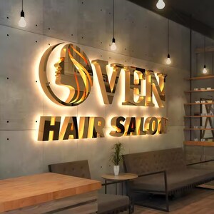 May include: A gold-toned, backlit 3D wall sign featuring a circular logo with a stylized profile of a face and the text OVEN HAIR SALON. The letters and logo are mounted on a gray concrete wall with visible dark pinholes and warm lighting.