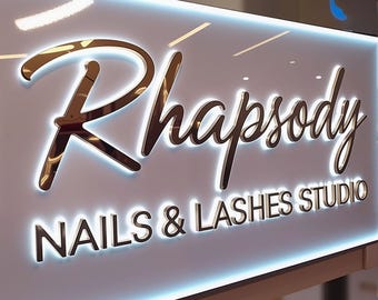 May include: A sign for "Rhapsody Nails & Lashes Studio" with gold lettering and a white background. The words "NAILS & LASHES STUDIO" are in a smaller font below "Rhapsody". The sign is illuminated with a soft, white light.
