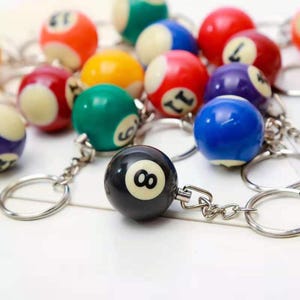 May include: A selection of billiard ball keyrings in various colours. The keyrings feature numbered balls, including a black "8" ball. The balls are attached to silver keyrings, creating a fun accessory.