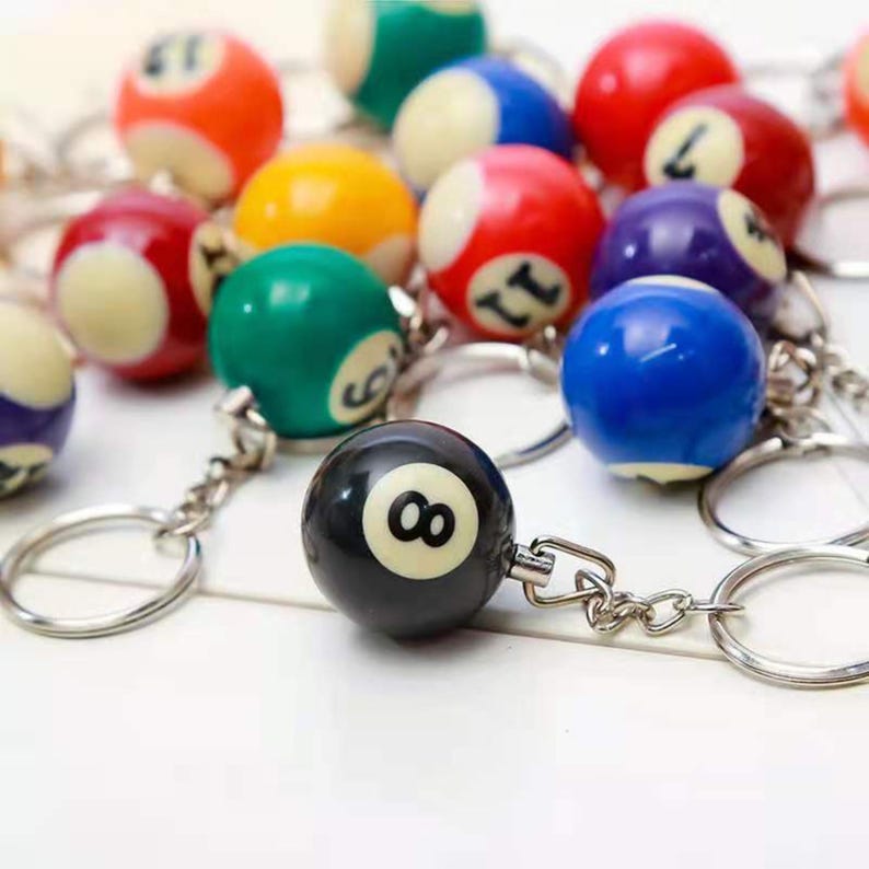 May include: A selection of billiard ball keyrings in various colours. The keyrings feature numbered balls, including a black "8" ball. The balls are attached to silver keyrings, creating a fun accessory.
