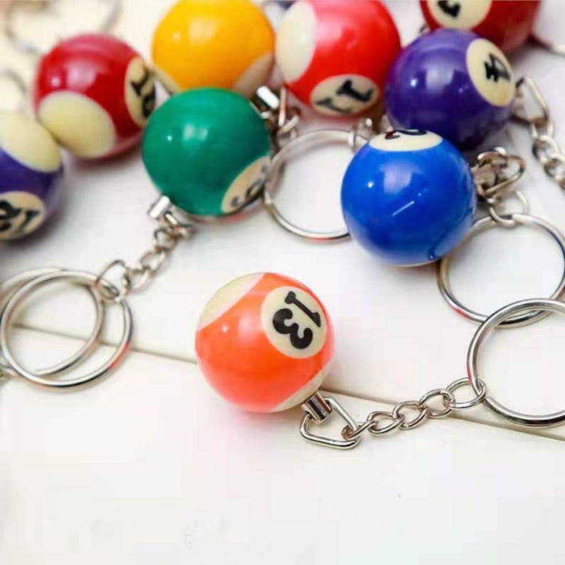 May include: A selection of billiard ball keyrings. Each keyring features a small, numbered billiard ball in various colours, including red, yellow, green, and blue. The keyrings are attached to silver-coloured metal rings and chains.