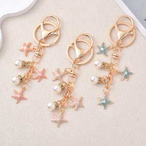 May include: Three gold-coloured keychains with lobster claw clasps. Each keychain features a cluster of starfish charms in pink, white, and blue, along with faux pearls and small rhinestone accents. The keychains are designed with a nautical theme.
