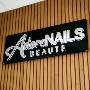 May include: A rectangular black sign with raised, metallic silver lettering that reads AdoreNAILS BEAUTE. The sign features a script font for the word Adore and a bold, sans-serif font for NAILS and BEAUTE, mounted on a vertical wood slat wall.