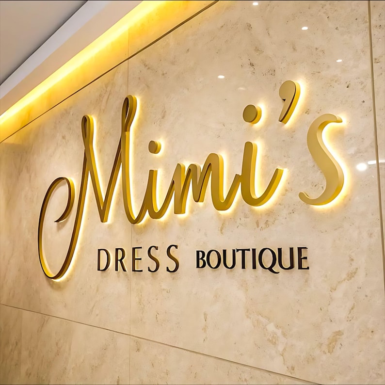 May include: A wall-mounted sign featuring the gold-colored, three-dimensional script text Mimi's with backlighting, positioned above the black, capitalized sans-serif text DRESS BOUTIQUE, all set against a polished, light beige marble-patterned wall.