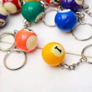 May include: A selection of colourful billiard ball keychains. The keychains feature various colours, including orange, yellow, green, and blue, each with a white circle and a number. The keychains are attached to silver key rings.
