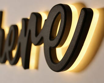 May include: A black, cursive-style word, "here", is mounted on a white wall. The letters are thick and have a textured appearance. A warm, yellow light emanates from behind the letters, creating a glowing effect. The sign is a decorative wall accent.