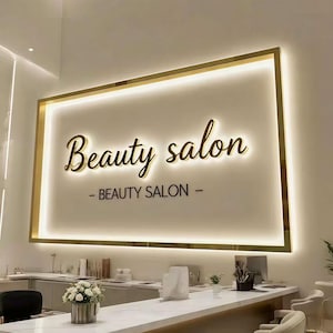 May include: A rectangular wall sign with a gold frame and integrated backlighting. The sign features the cursive text Beauty salon and the capitalized text BEAUTY SALON below it. The design is mounted on a plain wall above a white counter.