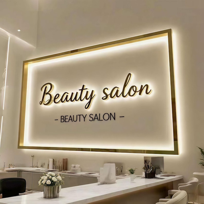May include: A rectangular wall sign with a gold frame and integrated backlighting. The sign features the cursive text Beauty salon and the capitalized text BEAUTY SALON below it. The design is mounted on a plain wall above a white counter.