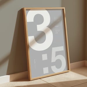 Könnte beinhalten: Ein gerahmtes Poster mit hellgrauem Hintergrund und weißen Zahlen "3:5" darauf gedruckt. Das Poster befindet sich in einem Holzrahmen und lehnt an einer Wand.