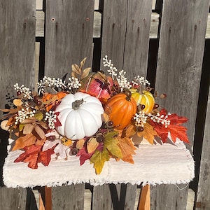 May include: A fall-themed centerpiece featuring a white pumpkin, orange and yellow pumpkins, and colorful autumn leaves. The arrangement includes decorative berries and is set on a cream-colored, fringed surface.