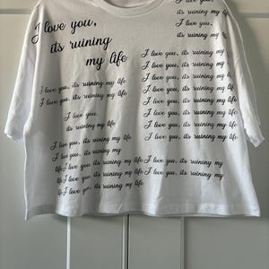 Peut inclure: T-shirt blanc avec l'inscription noire "I love you, it's ruining my life" répétée plusieurs fois. Le t-shirt est suspendu à un cintre noir. Le texte est répété sur le t-shirt.