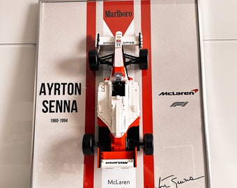 May include: Framed artwork featuring a McLaren MP4/4 race car model. The car is white and orange, set against a gray background with red stripes and the Marlboro logo. The frame includes the name "Ayrton Senna" and his birth and death years, along with the McLaren logo and signature.