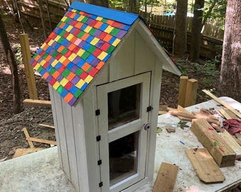Puede incluir: Una caja de intercambio de libros de madera blanca con un techo de cuadros de colores. La caja tiene una puerta de vidrio y una pequeña manija de metal. La caja está sentada en una mesa de madera con algunos trozos de madera cerca.