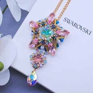 Op de afbeelding: Een gouden ketting met een roze, blauwe en groene stervormige hanger en een traanvormig kristal. De hanger is gemaakt van Swarovski-kristallen en kralen. De ketting ligt op een witte achtergrond met het woord "SWAROVSKI" erop gedrukt.