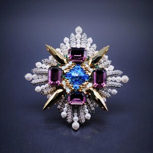 May include: An elaborate brooch featuring a central blue gemstone, framed by purple and gold elements. The design incorporates white pearls and clear crystals, giving a luxurious and ornate look. The brooch is set against a dark background.