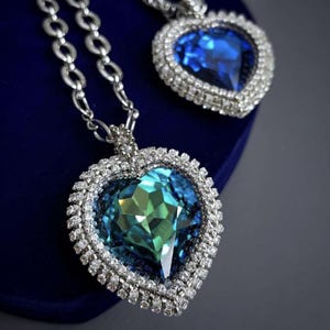 May include: Two silver heart-shaped pendants with blue crystal stones. The larger pendant has a green hue in the center of the stone.