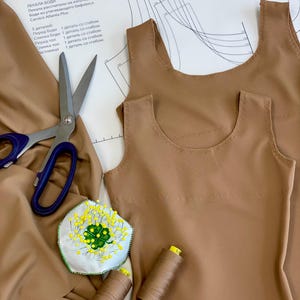 以下が含まれることがあります： 白い背景に茶色の布地のボディスのパターンをクローズアップした写真。パターンは、丸首のノースリーブボディス用です。茶色の糸の糸巻きがいくつかと、緑のクローバー模様の針山があります。