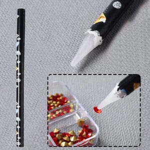 May include: A black rhinestone applicator tool with a white tip, used for picking up and placing small, red and gold rhinestones. The tool is decorated with silver and gold accents. A small container holds the rhinestones.