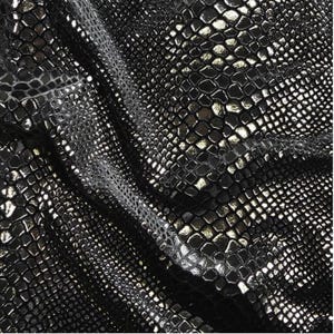 Könnte beinhalten: Nahaufnahme eines schwarzen Stoffes mit Schlangenmuster. Das Material hat ein glänzendes, metallisches Aussehen mit silbernen und goldenen Akzenten. Die Textur ist detailliert und der Stoff ist leicht zerknittert.