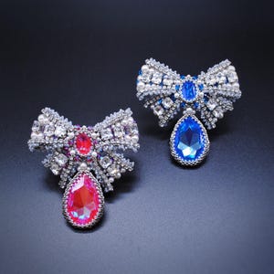 Könnte beinhalten: Zwei Perlenbroschen in Form von Schleifen mit tropfenförmigen Kristallen. Eine Brosche hat einen blauen Kristall und die andere einen rosa Kristall.