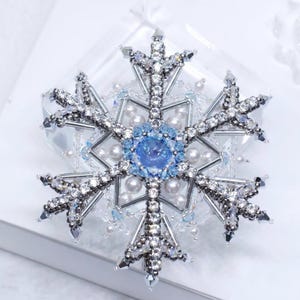 May include: A detailed, handcrafted snowflake brooch. The brooch features a central blue gemstone surrounded by clear and blue crystals, pearls, and silver-toned metal. The snowflake design is intricate and delicate, with each point adorned with sparkling accents.