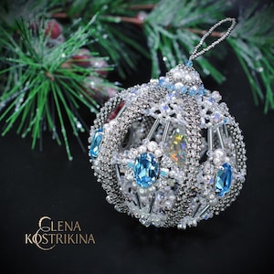 Puede incluir: Un adorno navideño artesanal con un diseño de cuentas. El adorno presenta un exterior de cuentas plateadas y blancas, adornado con piedras preciosas azules y perlas. Se suspende mediante un lazo plateado. El fondo incluye agujas de pino y el texto "GLENA KOSTRIKINA".