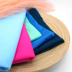 May include: A stack of five fabric swatches in different colors, including light blue, pink, teal, blue, and dark blue. The swatches are arranged on a wooden plate.