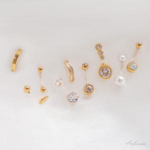 May include: A collection of gold-toned earrings and body jewelry, including hoops, studs, and dangling pieces. Some pieces feature clear gemstones and pearls, arranged on a white, textured surface. The jewelry is displayed in a studio setting with soft lighting.