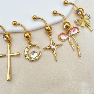 May include: Five gold belly button rings with different charms. The charms include a cross, a crescent moon, a star, and two bows with rhinestones.
