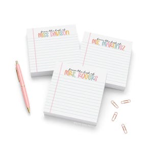 May include: Three square white sticky note pads with horizontal lines and a pink margin. Each pad features the text "from the desk of" followed by a name in colorful, rounded lettering. A light pink ballpoint pen and four rose gold paper clips.