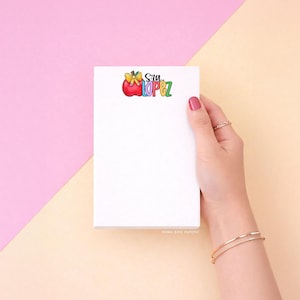 May include: A rectangular white notepad featuring a graphic at the top with a red apple, a yellow bow, and the text Sra. LOPEZ in colorful, stylized lettering. The notepad is blank below the graphic, providing space for notes and writing tasks.