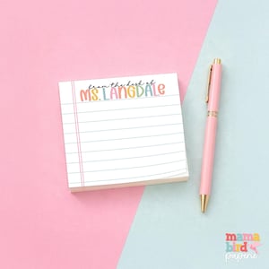 May include: A square notepad with horizontal blue lines and a pink vertical margin line. The top of the notepad features the text "from the desk of MS. LANGDALE" in colorful, rounded letters. A light pink ballpoint pen with gold accents is placed beside it.