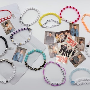 May include: A collection of beaded bracelets in various colors, including red, white, black, yellow, pink, and blue. Some bracelets feature letter beads spelling out phrases like "Farmhouse Fixer" and "A Love Like This." The image also includes small cards.