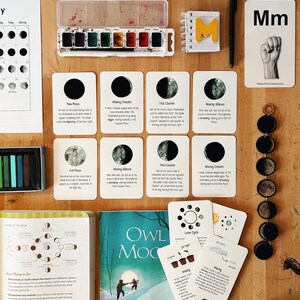 Pode incluir: Um conjunto de flashcards coloridos que ilustram as fases da lua com descrições. Os cartões estão dispostos sobre uma mesa de madeira com um livro, um conjunto de tintas de aquarela e um conjunto de lápis pretos e brancos. Os cartões são etiquetados com os nomes das fases da lua, como "Lua nova", "Lua crescente", "Quarto crescente", "Lua gibosa crescente", "Lua cheia", "Lua gibosa minguante", "Quarto minguante" e "Lua minguante".