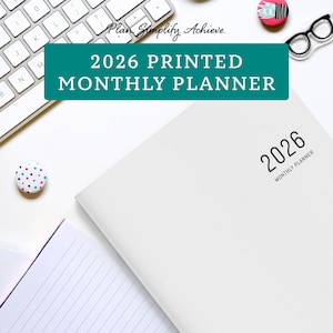 May include: A white 2026 monthly planner with the text "2026 MONTHLY PLANNER" printed on the cover. The image also includes a keyboard, decorative pins, and the text "2026 PRINTED MONTHLY PLANNER".