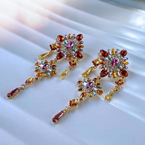May include: A pair of gold-tone dangle earrings featuring a cluster of multi-colored crystals in shades of pink, orange, yellow, and light blue. The design includes a square-shaped top section with dangling crystal accents and teardrop stones.