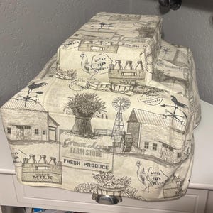 Puede incluir: Una cubierta de tostadora de tela beige y marrón con temática de granja. La cubierta presenta una ilustración en blanco y negro de una granja, botellas de leche y el texto "Green Acres Farm Store Fresh Produce".