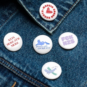 以下が含まれることがあります： 青いデニムジャケットに、デザインの異なる丸いピンが4つ付いている。ピンには、「KEIN HERZ, FÜR MERZ.」「PFOTEN WEG VON DER AFD」「FCK NZS」「FCK NZS」の文字が書かれている。